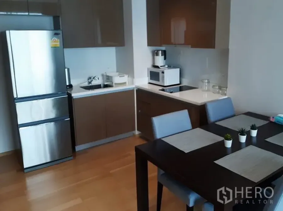 Condominium for rent in Khlong Toei, Bangkok - Modern condo kitchen with L-shaped cabinets, stainless fridge, cooktop, and a four-seat dining table.