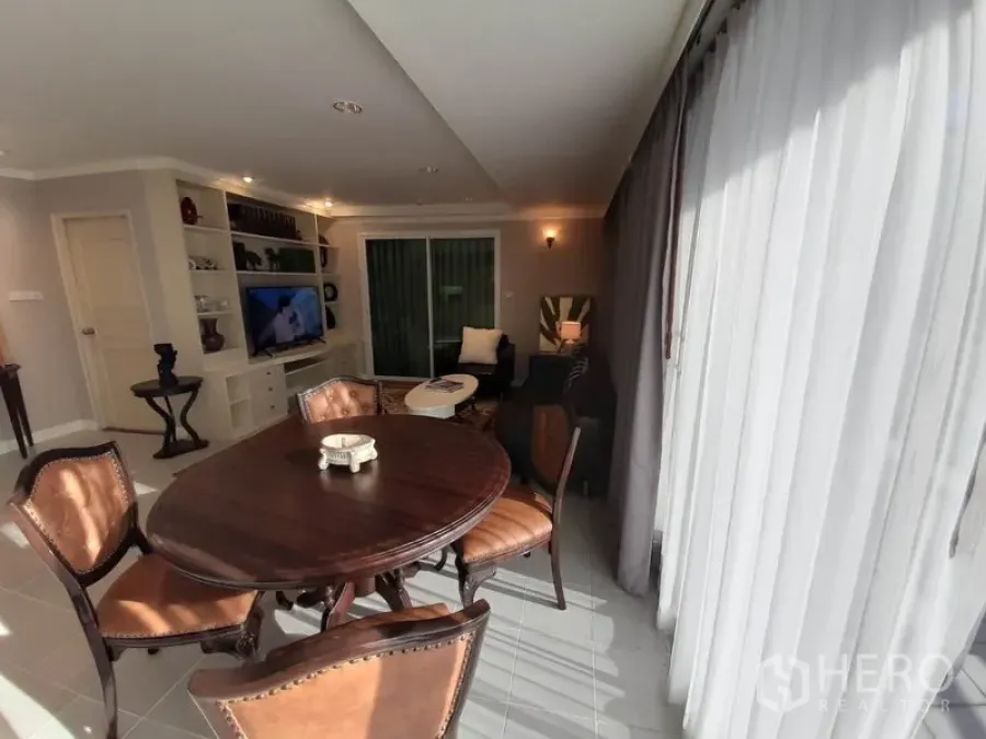 Condominium for rent or sale in Watthana, Bangkok - Sunlit living and dining area with a round wooden table, built-in shelving, TV, and sliding doors to the balcony behind sheer curtains.