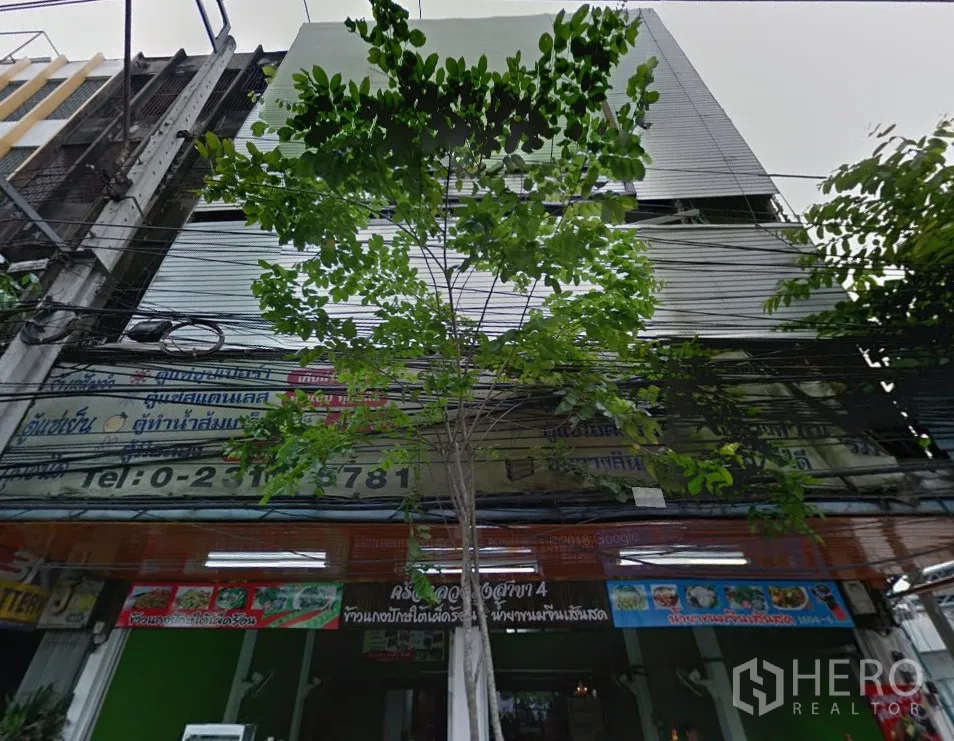 Shophouse for rent in Phra Khanong, Bangkok - Street‑level facade of the shophouse row with roller shutters and shop signage beneath leafy trees.