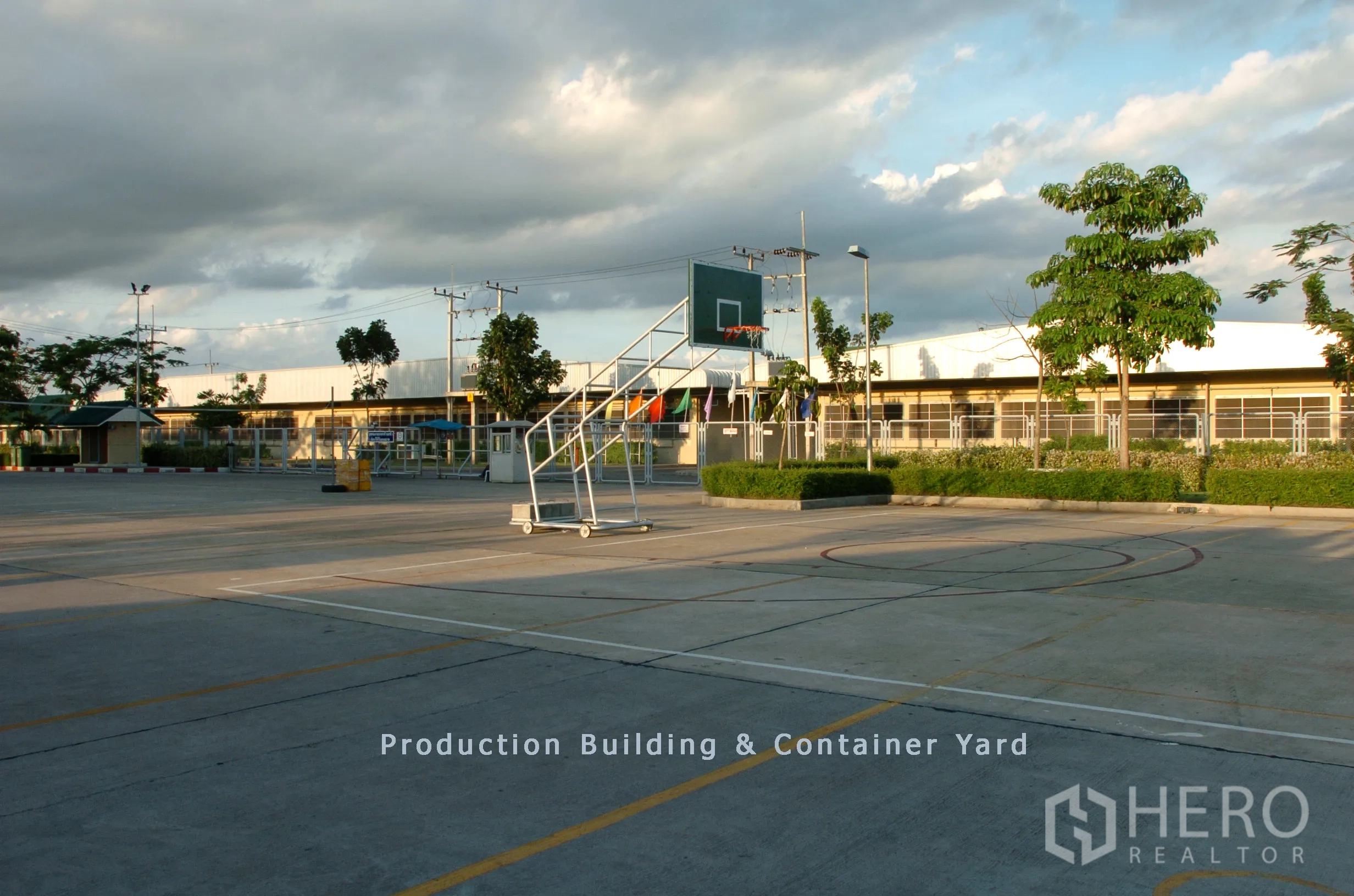 Factory for sale in Khao Chamao, Rayong - Wide container yard and production building with a movable basketball hoop, perimeter fence and greenery under a cloudy sky.