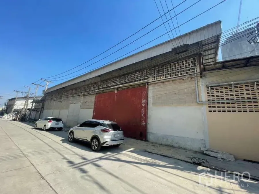 Warehouse for rent in Rat Burana, Bangkok - Long warehouse frontage with red gate and utility lines along a wide concrete access road.