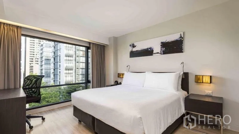房间 出租在Watthana, Bangkok - 卧室配大床、书桌与落地窗，俯瞰曼谷城市天际线。