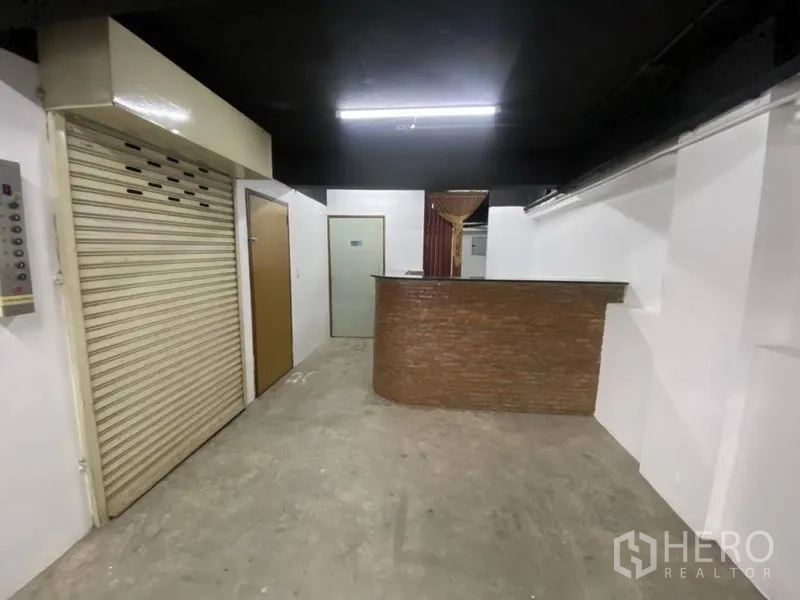Retail Space for rent in Bangkok - Retail interior with reception counter, roller shutter door, white walls, and concrete floor under fluorescent lighting.