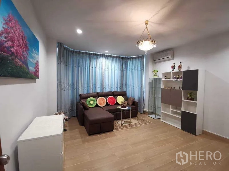 Detached House for rent in Prawet, Bangkok - Cozy living room with L‑shaped brown sofa, blue curtains, chandelier, air‑conditioning and display shelves.