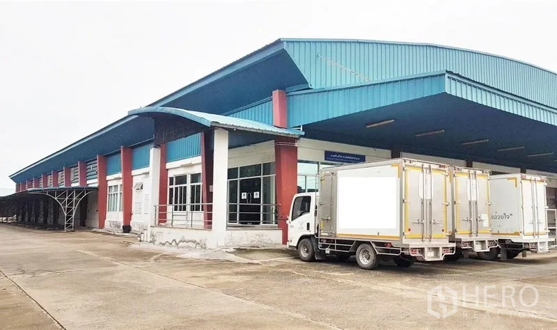 Factory for sale in Lam Luk Ka, Pathum Thani - Large factory with blue metal roof, glass-fronted office and dock‑high loading bays with trucks parked.