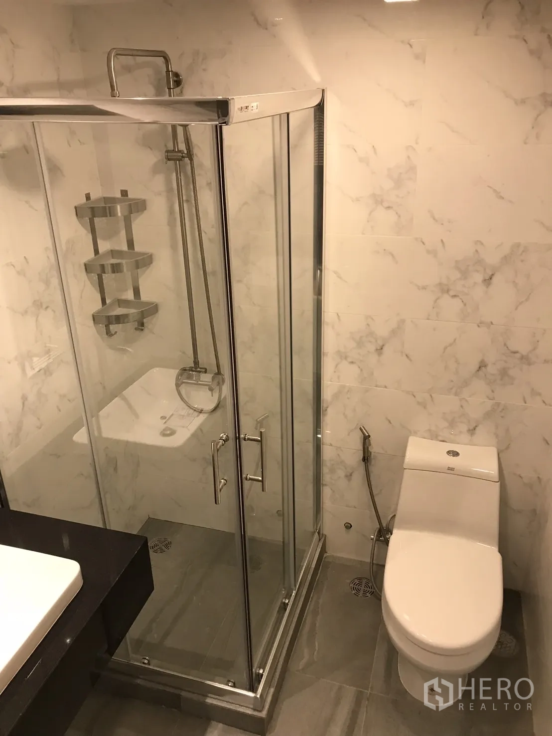 Condominium for rent or sale in Khlong Toei, Bangkok - Bathroom view showing glass shower enclosure, toilet and gray floor tiles with marble walls.