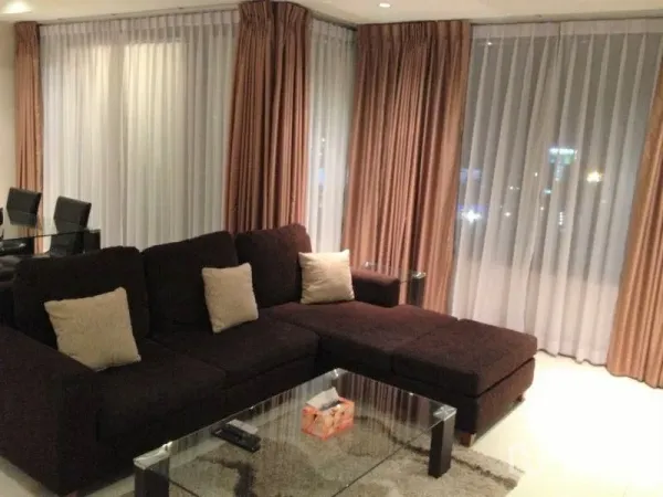 Condominium for rent or sale in Khlong Toei, Bangkok - Living room featuring a dark L-shaped sofa, glass coffee table, and large windows with drapes.