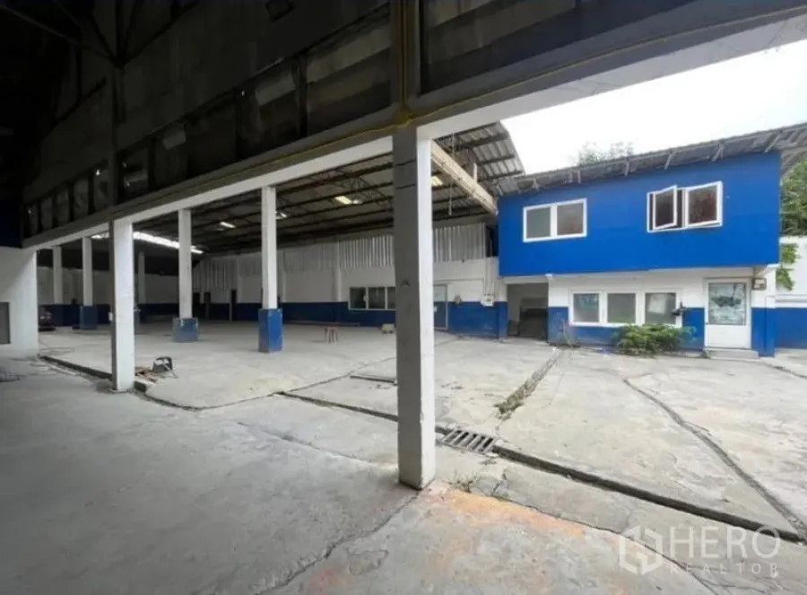 Warehouse for rent in Phra Khanong, Bangkok - Covered loading area with pillars beside a blue two-storey office and wide concrete apron.