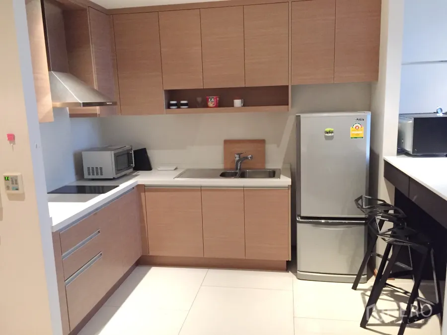 Condominium for rent in Khlong Toei, Bangkok - Modern L-shaped kitchen with wood cabinets, induction hob, stainless hood, microwave and fridge in a Bangkok condo.