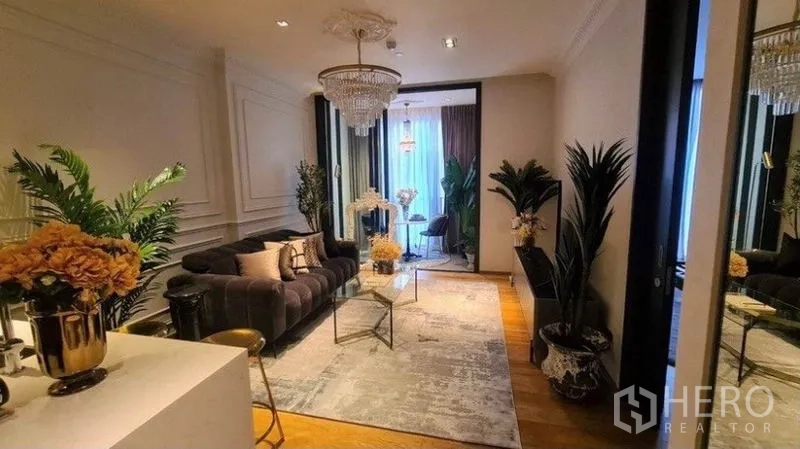 Condominium for rent in Khlong Toei, Bangkok - Elegant living room with chandelier, velvet sofa, glass coffee table, wall molding and indoor plants leading to a bright windowed nook.