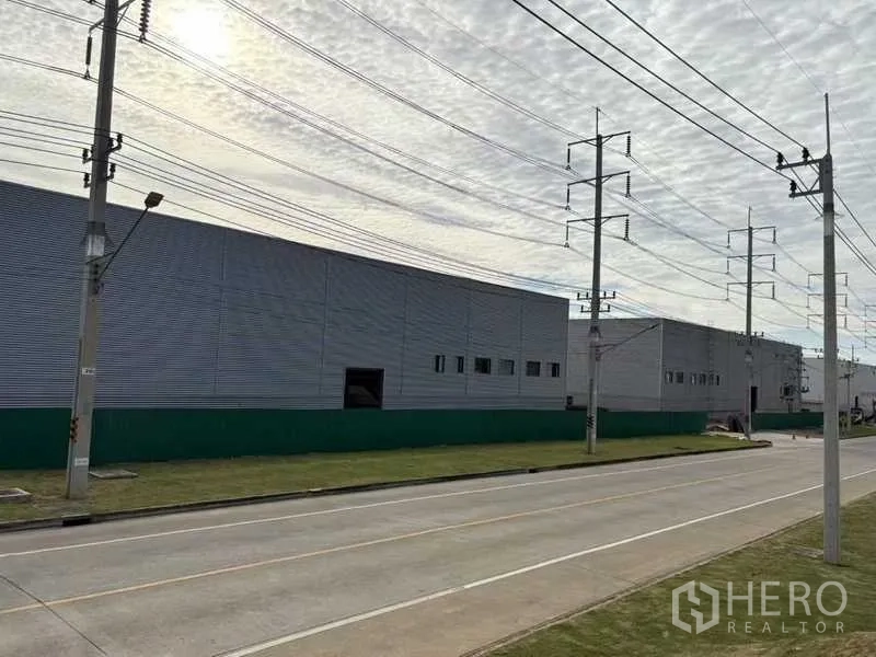 Factory for rent in Rayong - Side view of an industrial warehouse beside a paved road and utility poles in Rayong.