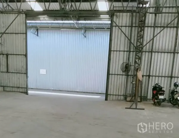 Warehouse for rent in Khlong Luang, Pathum Thani - Interior of warehouse showing open sliding doors, steel trusses, concrete floor and a ventilation fan with wide vehicle entry.