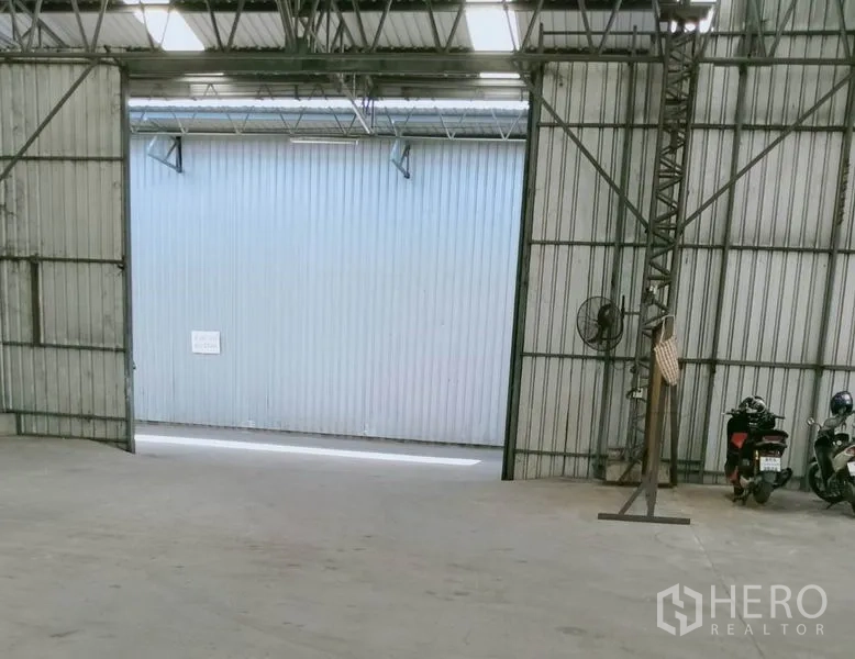 Warehouse for rent in Khlong Luang, Pathum Thani - Interior of warehouse showing open sliding doors, steel trusses, concrete floor and a ventilation fan with wide vehicle entry.