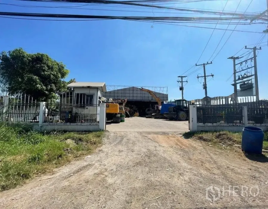 Warehouse for rent in Wang Noi, Phra Nakhon Si Ayutthaya - Gated warehouse entrance with guardhouse, wide driveway to a steel-roof shed and yard with heavy machinery, power poles overhead under a clear blue sky.
