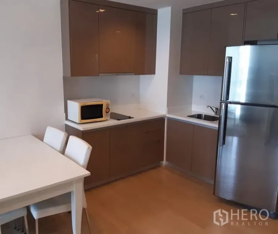 Condominium for rent in Khlong Toei, Bangkok - Modern L-shaped kitchen with glossy cabinets, stainless refrigerator, microwave and a dining table in a Bangkok condo.