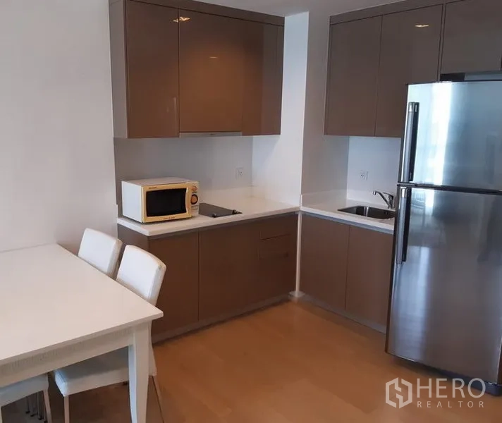 Condominium for rent in Khlong Toei, Bangkok - Modern L-shaped kitchen with glossy cabinets, stainless refrigerator, microwave and a dining table in a Bangkok condo.
