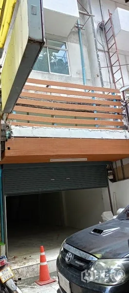 店屋 出租在Watthana, Bangkok - 店屋外立面，木质横条阳台与半开的金属卷闸门，旁有脚手架和停放车辆。
