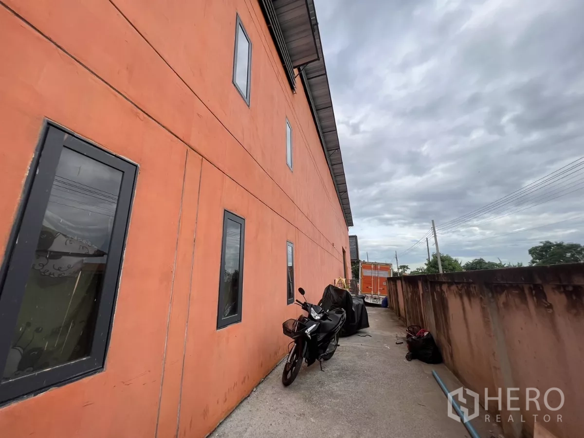Warehouse for rent or sale in Lat Krabang, Bangkok - Long side walkway beside the orange wall with narrow windows and a motorcycle under roof eaves.