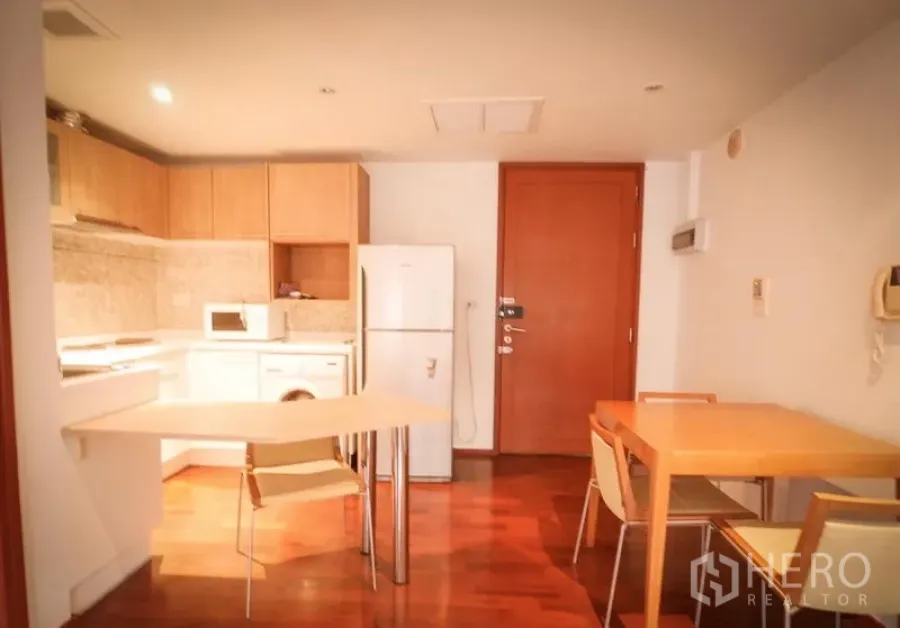 Condominium for rent in Pathum Wan, Bangkok - Open-plan kitchen with wood cabinetry, fridge, microwave, washing machine and dining tables on polished timber floors.