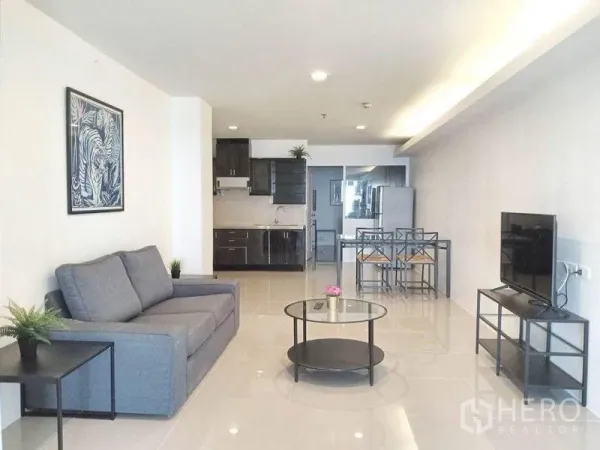 Condominium for rent in Khlong Toei, Bangkok - Open-plan living area with grey sofa, round coffee table, TV stand, dining set and kitchenette in bright Sukhumvit condo.