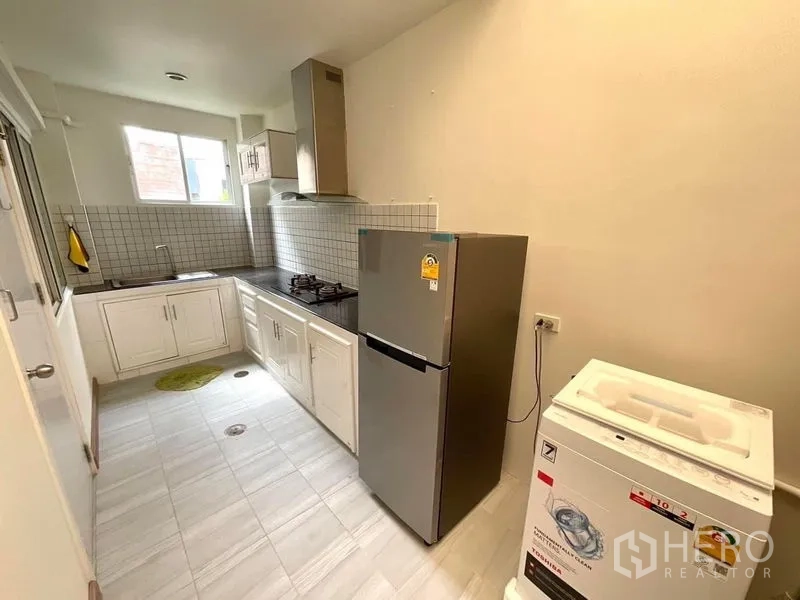 Townhouse for rent in Bang Phli, Samut Prakan - Fitted L-shaped kitchen with gas hob, hood, tiled backsplash, fridge and washing machine.