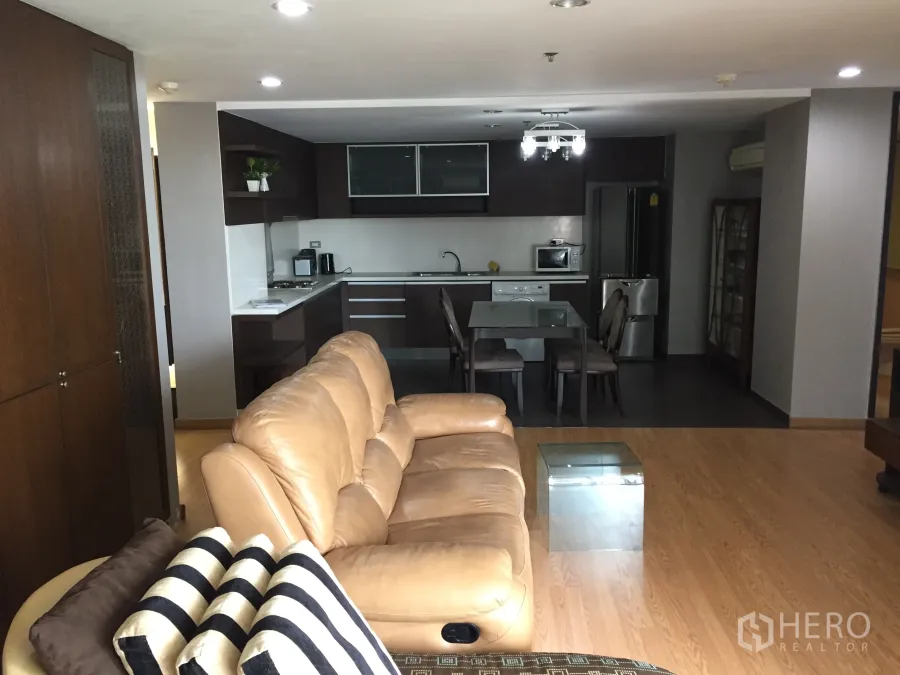 Condominium for rent in Watthana, Bangkok - Open-plan living room with tan leather sofa, glass dining table, and a fitted dark-wood kitchen with appliances in a Bangkok condo.
