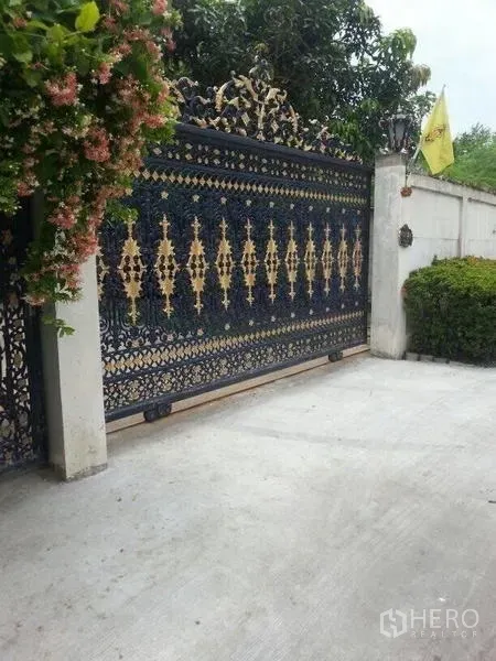 Detached House for rent in Bang Na, Bangkok - Ornate wrought-iron sliding entrance gate with gold accents beside landscaped shrubs.
