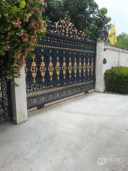 Detached House for rent in Bang Na, Bangkok - Ornate wrought-iron sliding entrance gate with gold accents beside landscaped shrubs.