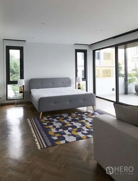 Townhouse for sale in Watthana, Bangkok - Bright master bedroom with large windows opening to a terrace, herringbone wood floor and a gray upholstered bed.