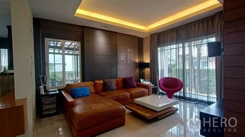 Detached House for sale in Prawet, Bangkok - Living room with L-shaped leather sofa, cove ceiling lights and wide glass doors to patio.