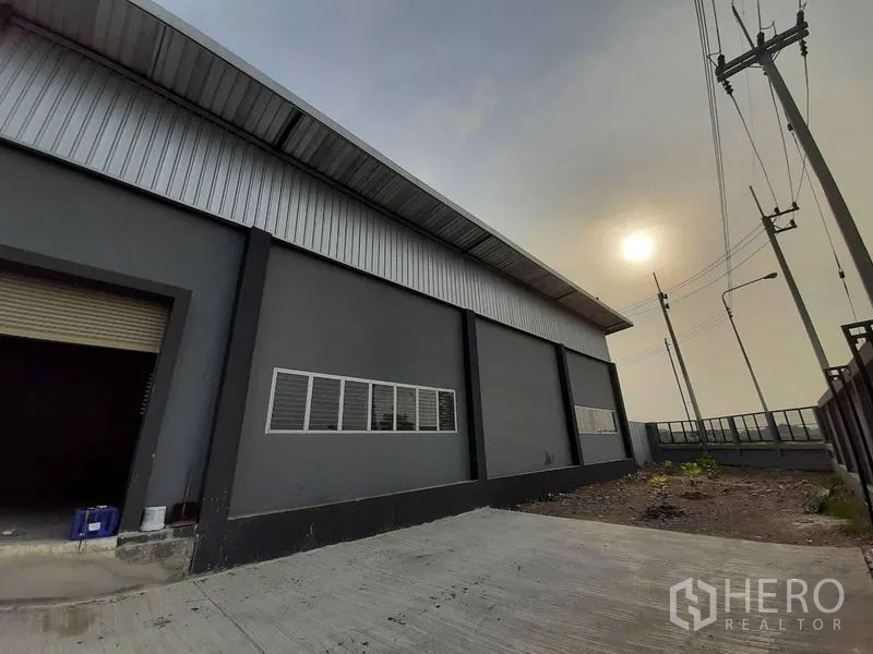 Warehouse for rent in Lam Luk Ka, Pathum Thani - Front exterior of a gray warehouse with concrete driveway, roller shutter door, and perimeter wall.