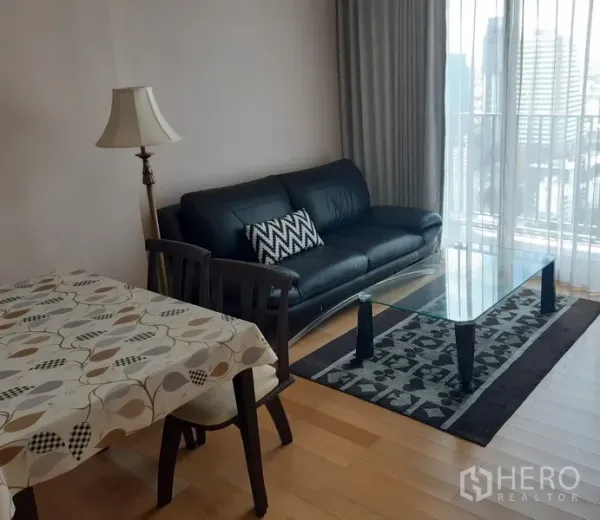 Condominium for rent in Khlong Toei, Bangkok - Bright living area with black leather sofa, glass coffee table, rug and floor-to-ceiling windows.
