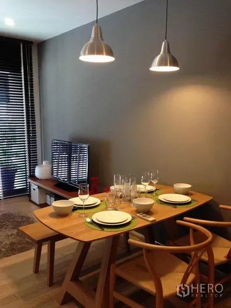 Condominium for rent in Watthana, Bangkok - Warm dining area with wooden table set for four, pendant lights, TV console and blinds by the balcony door.