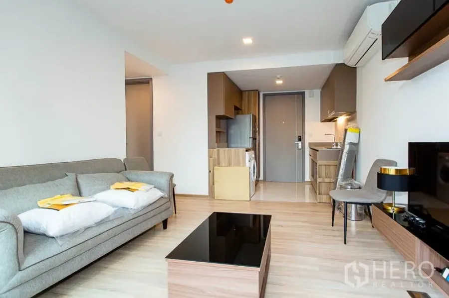 Condominium for rent in Watthana, Bangkok - Open-plan living room with gray sofa, wood floors, compact kitchen with fridge and washer, and TV unit.