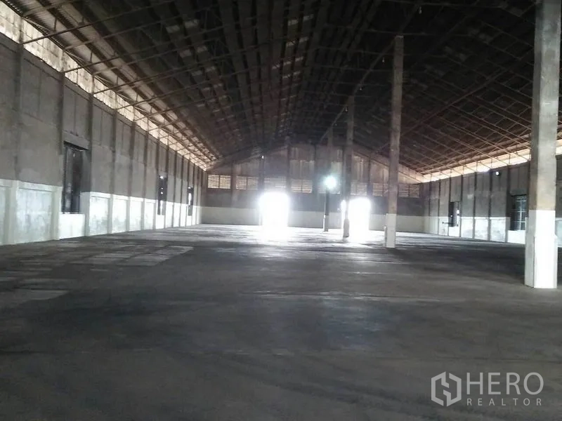Warehouse for rent in Pathum Thani - Spacious warehouse interior with high steel roof, concrete floor, central columns and bright light at loading doors.