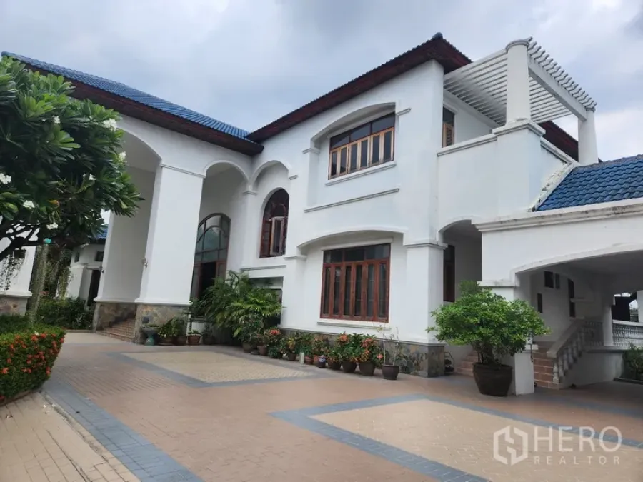 Detached House for rent in Prawet, Bangkok - Side exterior of the white villa with blue roof, covered walkway and potted greenery.