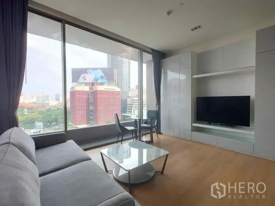 Condominium for rent in Bang Rak, Bangkok - Living area with TV wall unit, sofa and coffee table next to full-height windows facing city buildings.