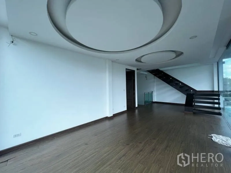 Detached House for sale in Lat Phrao, Bangkok - Spacious upper living room with wood floors, circular ceiling details, and open staircase by glass wall.