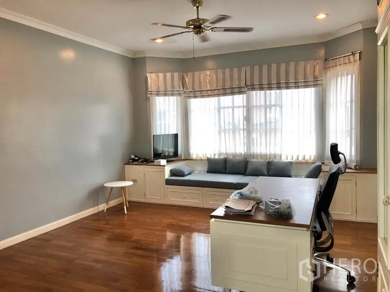 Detached House for rent in Mueang Samut Prakan, Samut Prakan - Spacious study or family room with wood floors, built‑in desk and bay‑window seat, ceiling fan and soft gray walls.