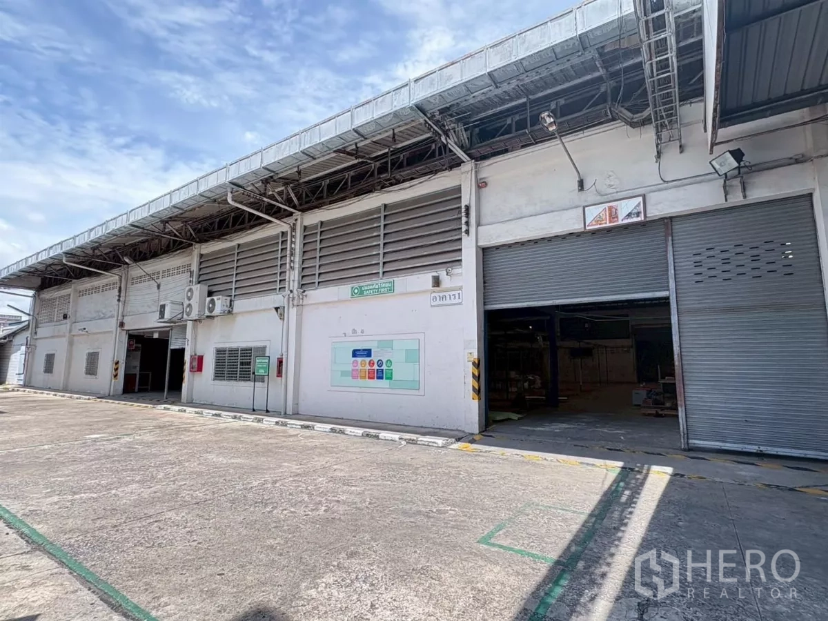 Factory for rent or sale in Bang Khun Thian, Bangkok - Wide exterior of an industrial factory with multiple roller shutters and a large driveway under a blue sky in Bang Khun Thian, Bangkok.