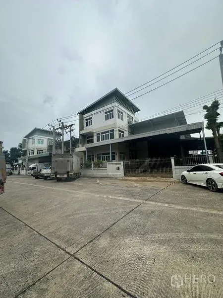 Warehouse for rent or sale in Lam Luk Ka, Pathum Thani - Street view of a modern three‑storey warehouse with gated loading bay and parked trucks in an industrial lane.