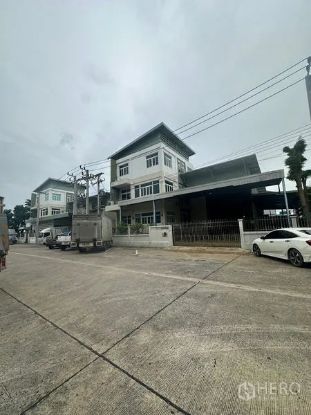 Warehouse for rent or sale in Lam Luk Ka, Pathum Thani - Street view of a modern three‑storey warehouse with gated loading bay and parked trucks in an industrial lane.