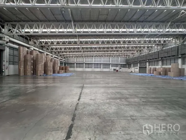 工厂 出租 - 宽阔仓库地面，钢桁架屋顶与采光带，一侧堆放纸卷。