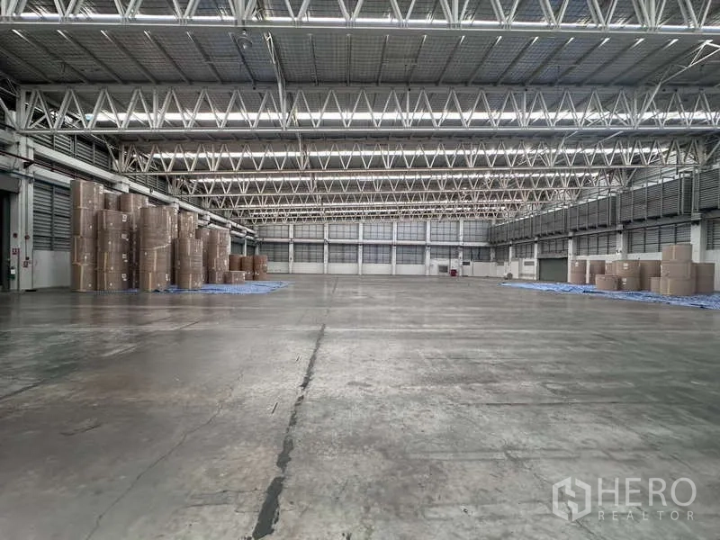 工厂 出租 - 宽阔仓库地面，钢桁架屋顶与采光带，一侧堆放纸卷。