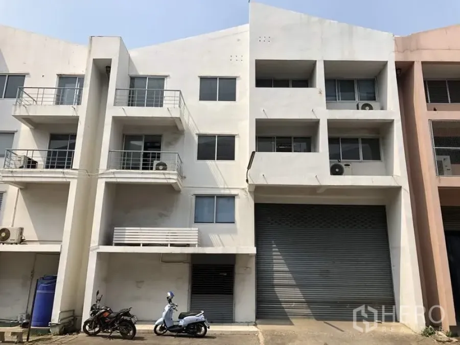 Factory for rent or sale in Khlong Luang, Pathum Thani - Multi-storey white factory-warehouse facade with large roller shutter, balconies and AC units in Khlong Luang.