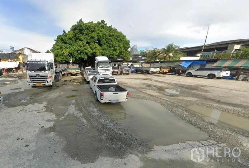Land for rent in Bang Na, Bangkok - Open gravel yard in Bang Na used for truck and pickup parking, with a large shade tree and roadside sheds.