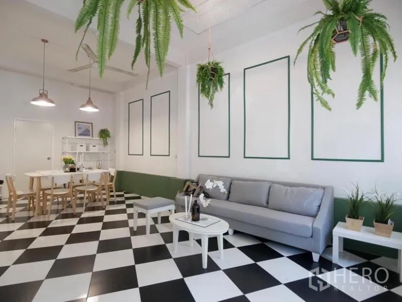 Townhouse for rent in Khlong Toei, Bangkok - Spacious lounge with a long gray sofa, pendant lights, decorative wall panels and checkered tiles leading to the dining corner.