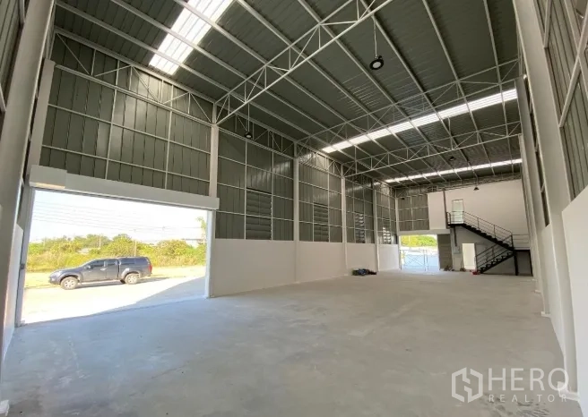 Warehouse for sale in Sai Noi, Nonthaburi - Spacious high-bay warehouse interior with steel truss roof, large roller shutters, smooth concrete floor, and mezzanine with stairs.