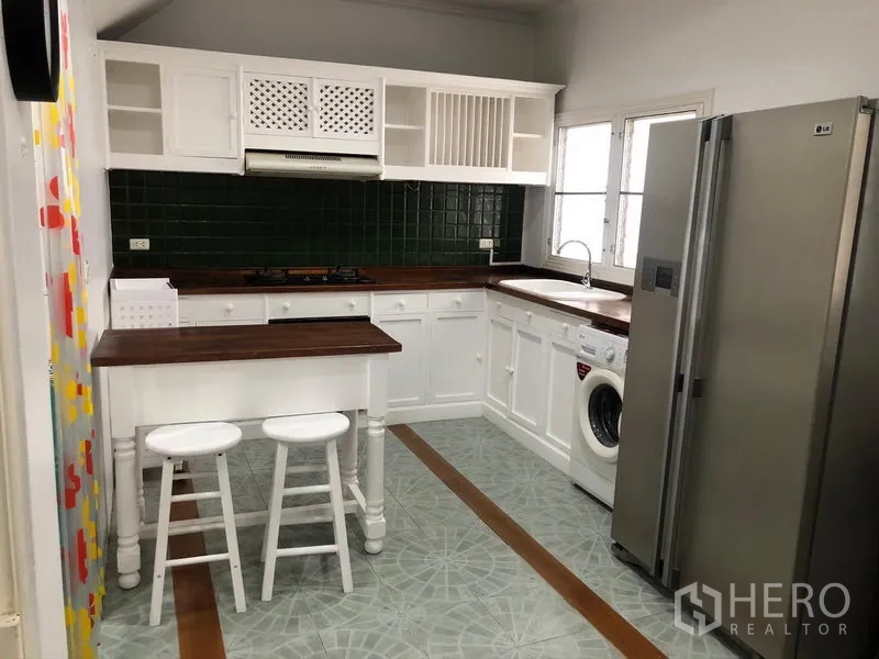 Townhouse for rent in Mueang Samut Prakan, Samut Prakan - Bright closed kitchen with white cabinetry, wood countertops, green tile backsplash, island with stools, fridge and washing machine.