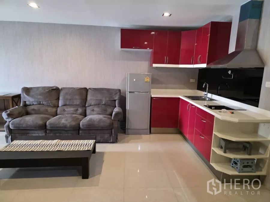 Condominium for rent in Khlong Toei, Bangkok - Open-plan living room with gray sofa and red L-shaped kitchen featuring stovetop, hood and refrigerator.